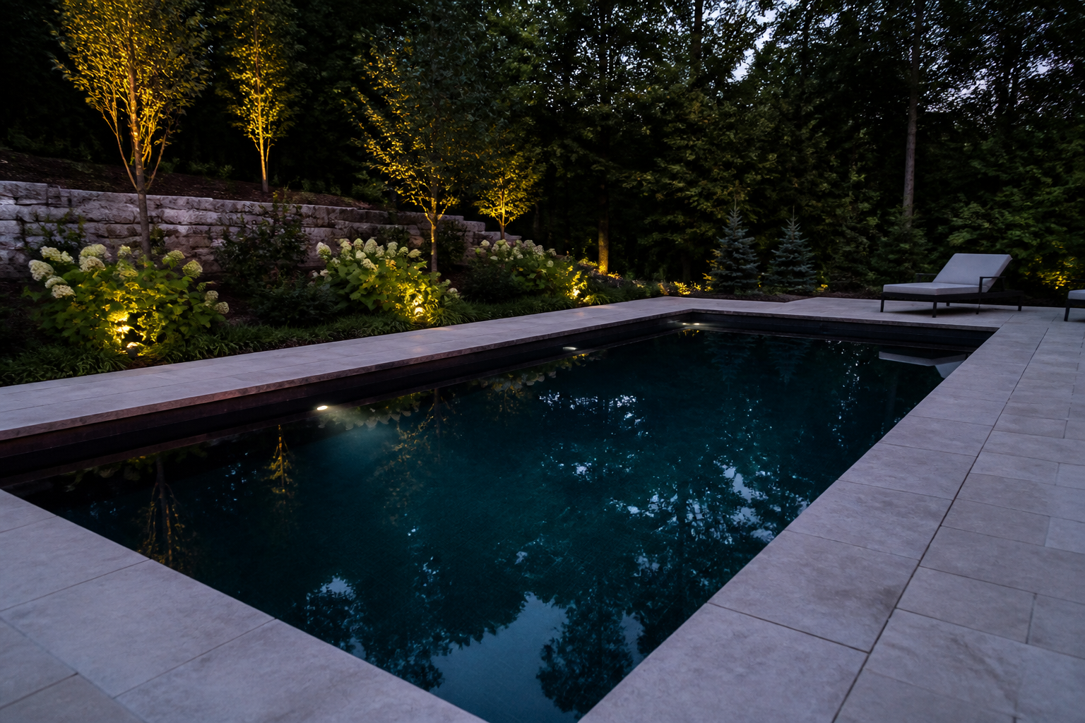A modern gunite swimming pool with a dark finish, surrounded by a light-coloured stone patio and gardens on an estate property at twilight.